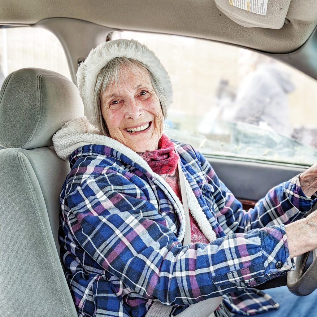 Donna, wearing a blue and purple plaid jacket and ivory hat, smiles at the camera from the driver's seat of her vehicle.